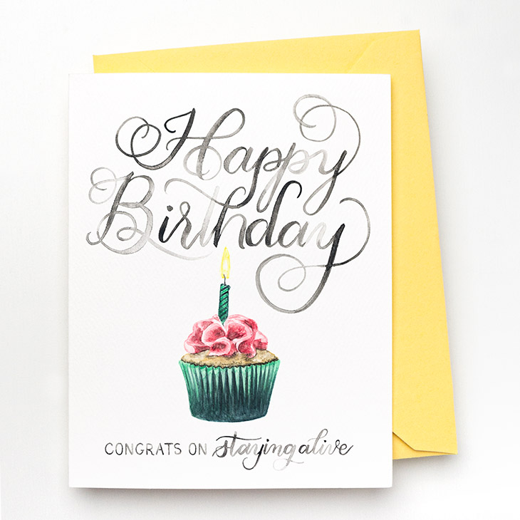 A hand-lettered watercolor card saying "Happy Birthday... congrats on staying alive" with a painted cupcake with one lit candle by CharmCat | charmcat.net