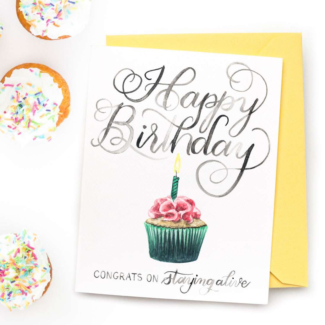 A hand-lettered watercolor card saying "Happy Birthday... congrats on staying alive" with a painted cupcake with one lit candle by CharmCat | charmcat.net